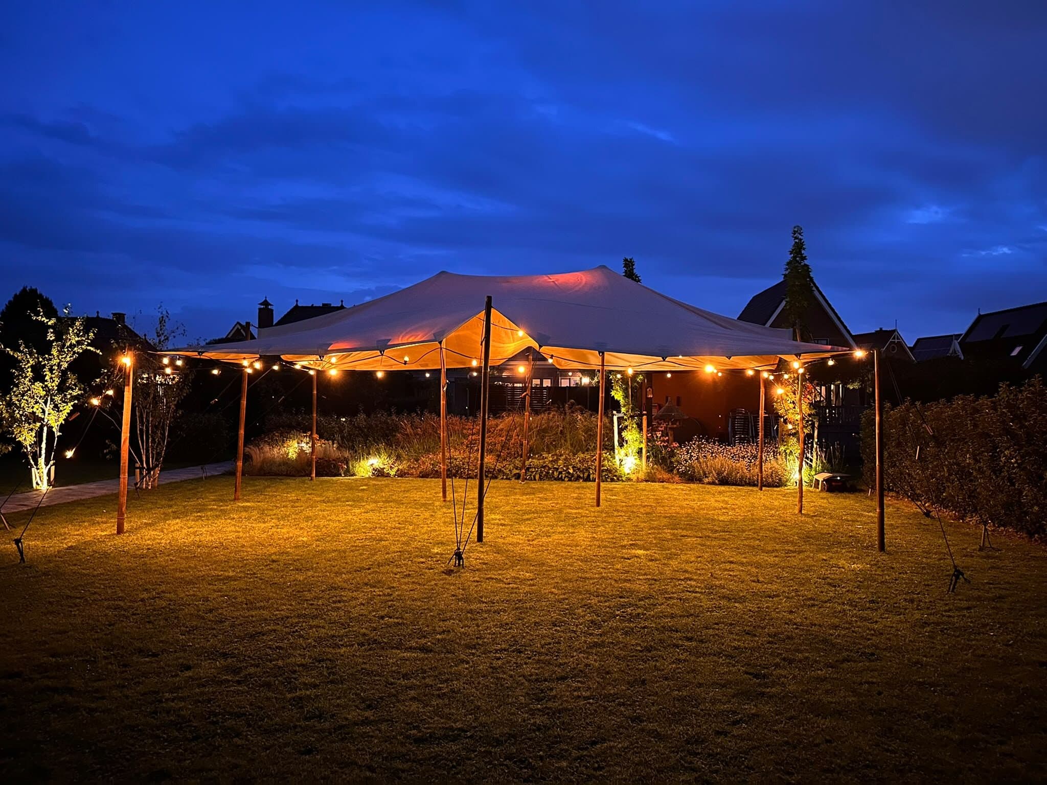 Beige stretchtent met brandende lampjes in een tuin bij avond