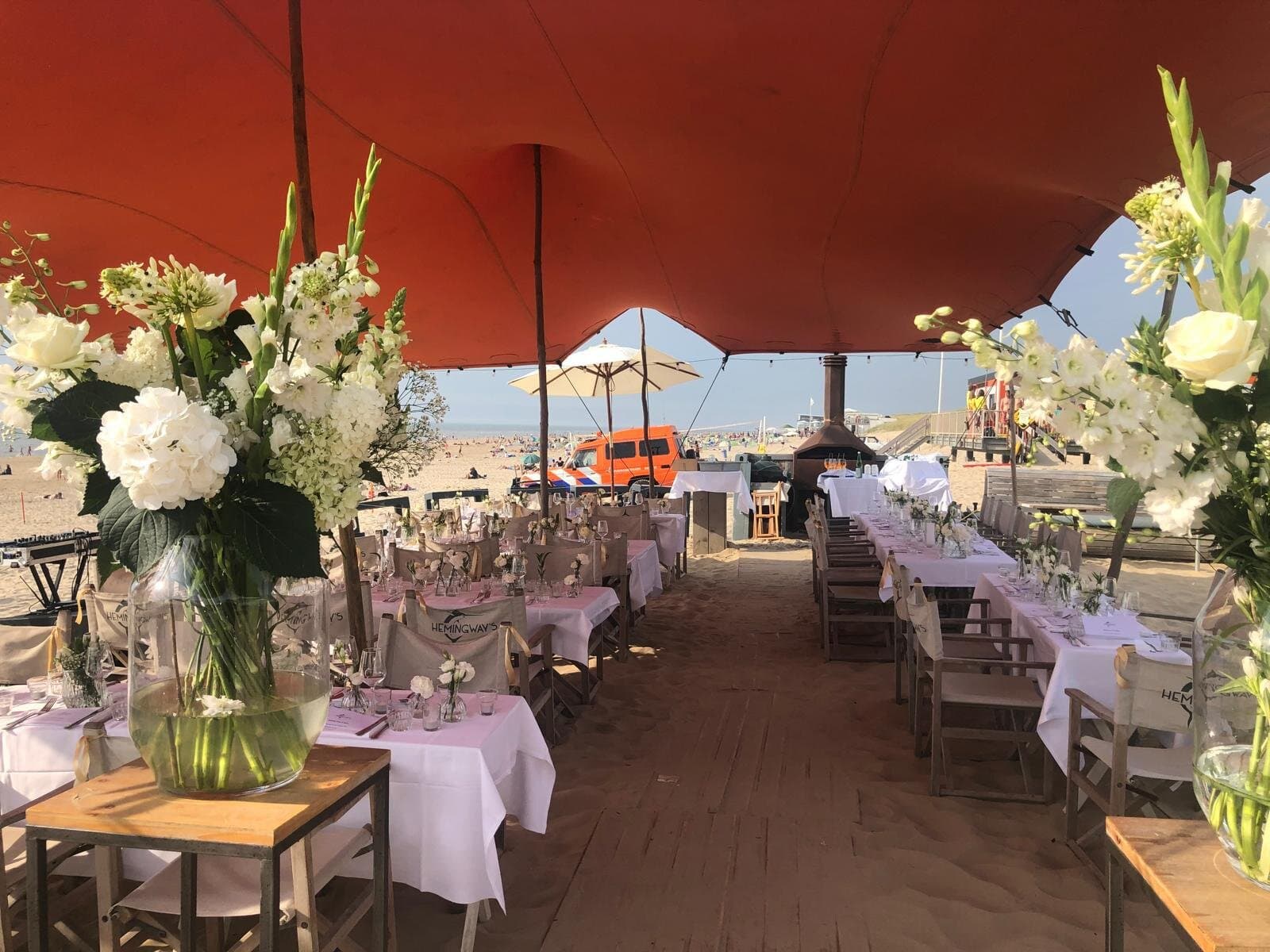 Witte bloemen op gedekte tafels onder een oranjerode stretchtent op het strand