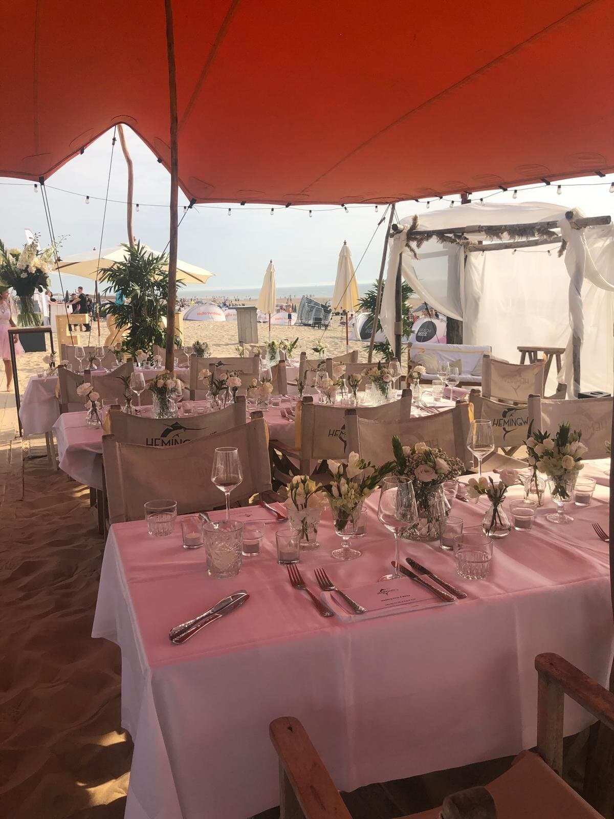 Gedekte tafels met witte kleden onder een oranjerode stretchtent op het strand