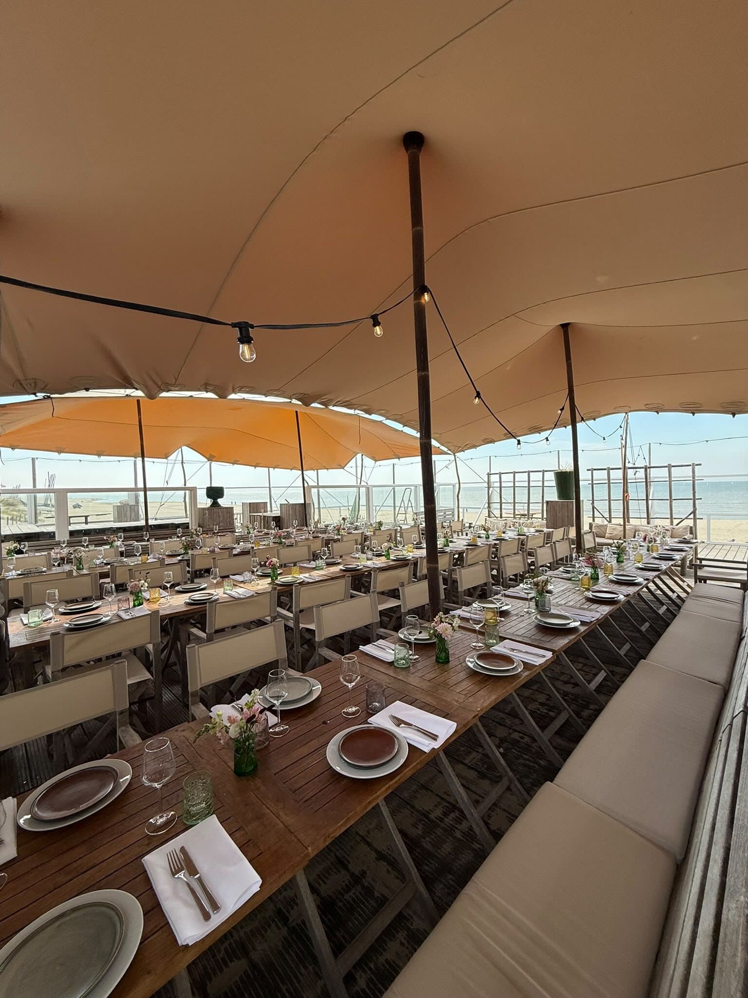 Gedekte tafels onder een beige en oranje stretchtent op een strandpaviljoen